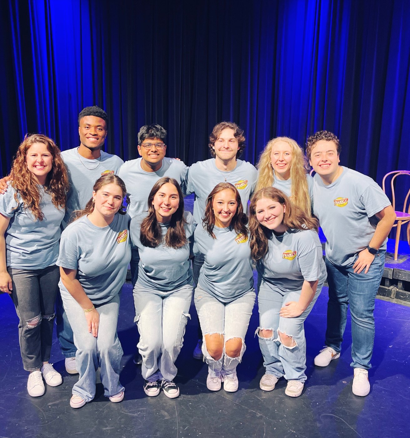 A troupe of smiling teen improv students on stage in matching shirts